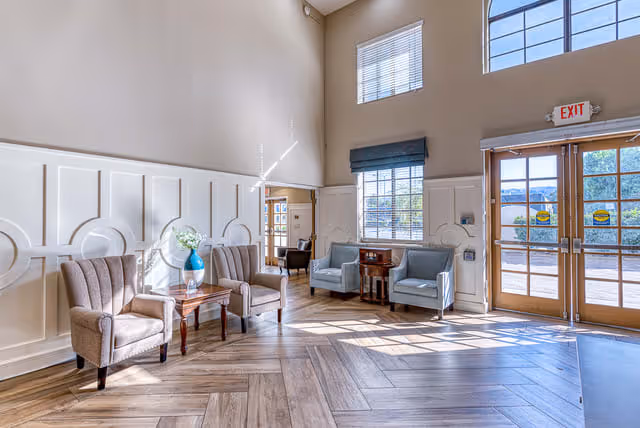 Sunlit senior living lobby with upholstered armchairs and side tables beneath tall windows and double glass doors.