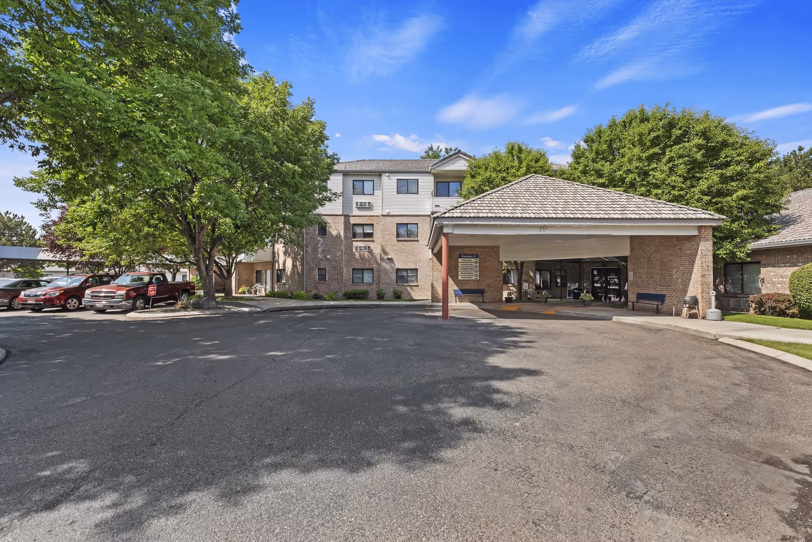 Exterior view of Karcher Senior Living facility showing a three-story building with a covered entrance driveway. There are several trees and parked cars in the parking area under a clear blue sky.