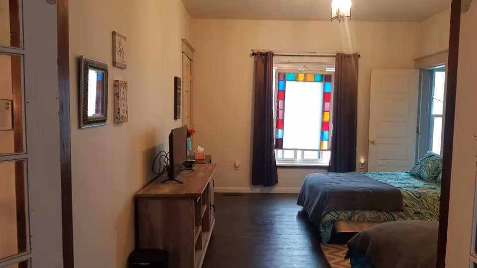 Interior view of a bedroom in an assisted living facility showing two beds with green and blue patterned bedding. There is a wooden TV stand with a television on it against the left wall. A window with colorful stained glass panels and dark curtains is at the far end of the room. The room has wooden flooring and light-colored walls with some framed artwork.
