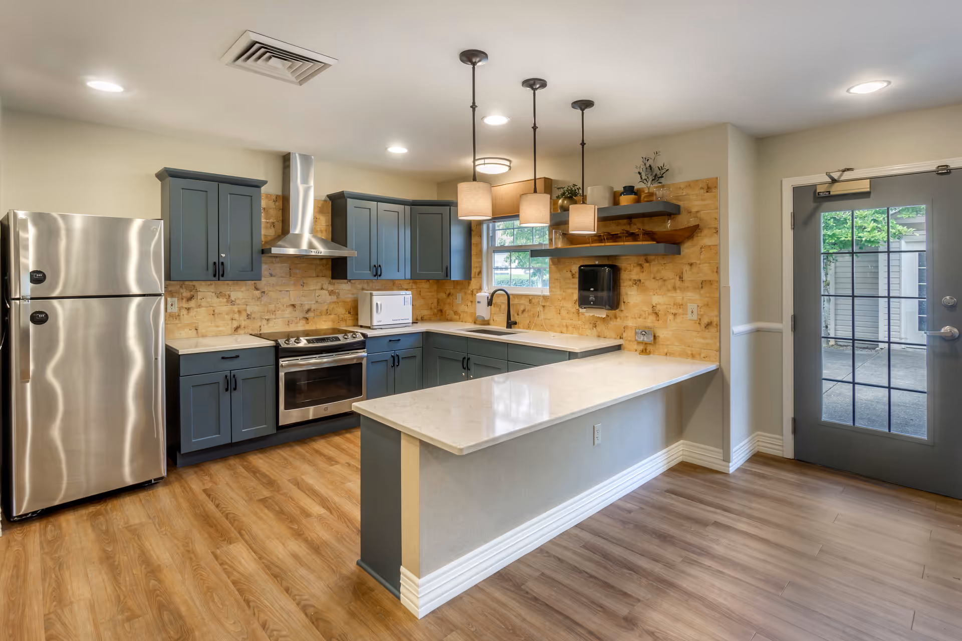 Modern kitchen with blue cabinets, stainless steel refrigerator and oven, a white countertop island with three pendant lights above, wooden flooring, and a door with glass panes leading outside.