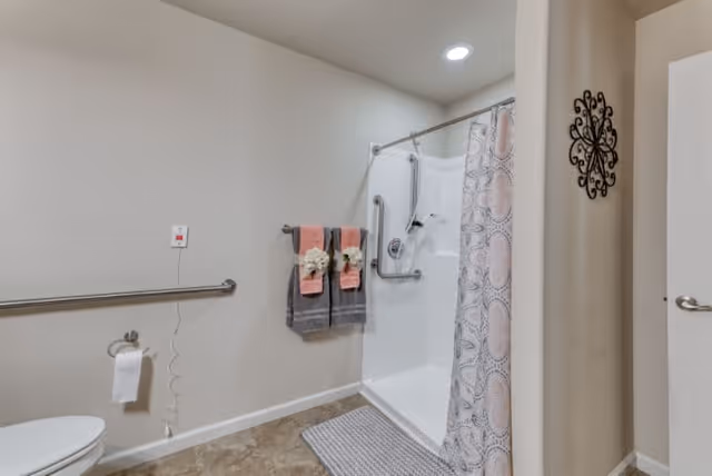 A clean and accessible bathroom with a white shower area featuring grab bars and a patterned shower curtain. Two sets of folded towels in pink and gray hang on a towel rack, each decorated with a white flower. A toilet paper holder with a roll of toilet paper is mounted on the wall next to the toilet. The floor is tiled, and a gray bath mat is placed in front of the shower. The walls are painted light beige, and a decorative black metal wall art piece is visible near the door.