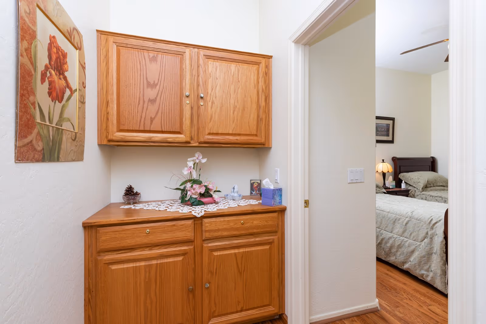 A hallway area with wooden cabinets mounted on the wall and below, decorated with a lace doily, a small flower arrangement, a pine cone in a glass bowl, a tissue box, and a small framed picture. To the right, an open doorway reveals a bedroom with a bed, nightstand, lamp, and framed picture on the wall. The floor is wooden and the walls are painted white.