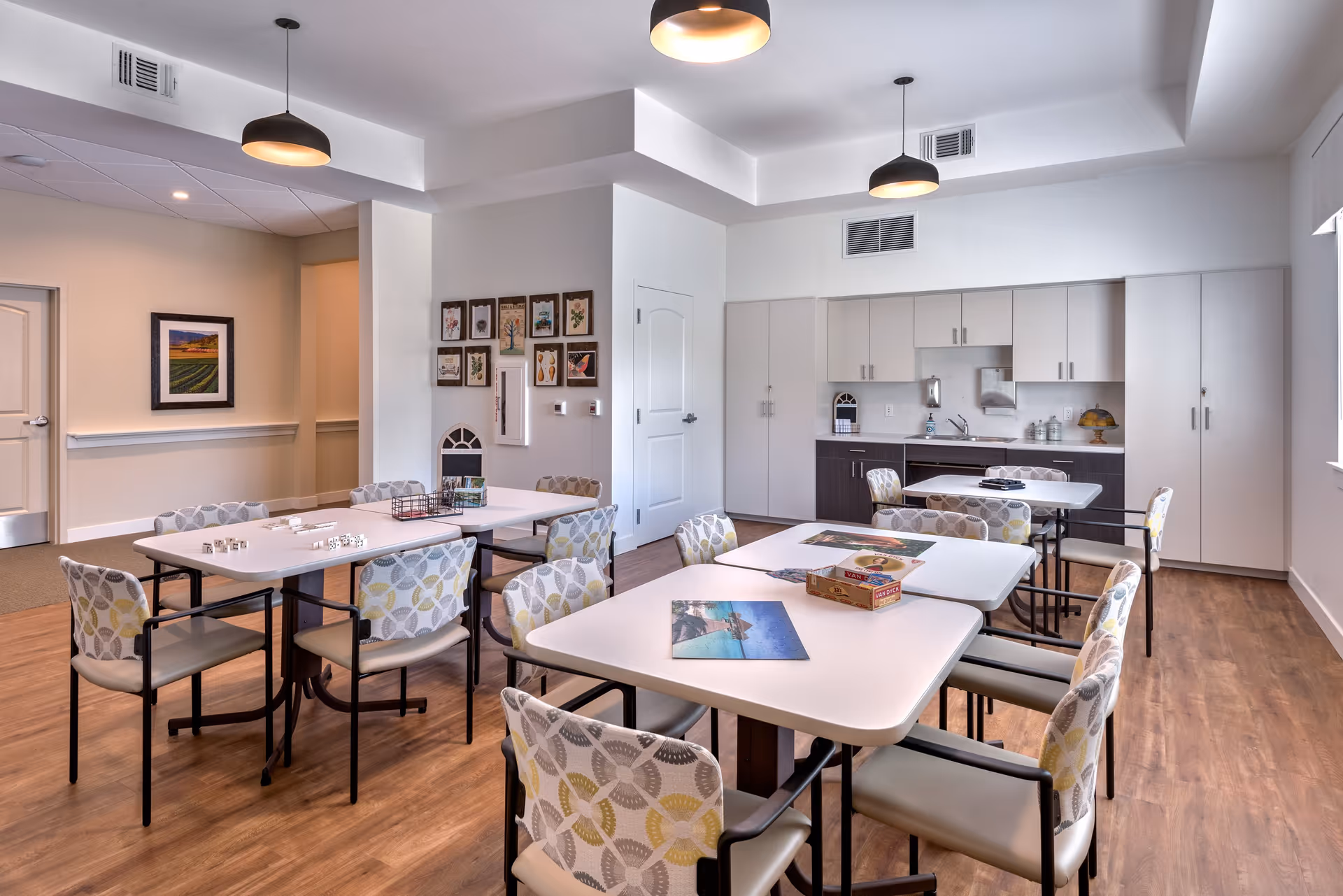 A bright and clean activity room in a senior living facility with several tables and chairs arranged for group activities. The tables have puzzles and board games on them. The room features wood flooring, white walls, modern ceiling lights, and a kitchenette area with cabinets and a sink in the background. Framed artwork decorates one wall.