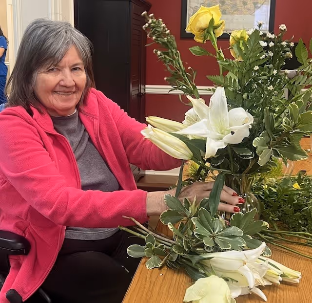 An elderly woman with gray hair wearing a pink jacket and gray shirt is sitting at a wooden table arranging a bouquet of flowers including white lilies and yellow roses. She is smiling and appears to be in a cozy indoor setting with red walls and a framed map on the wall behind her.