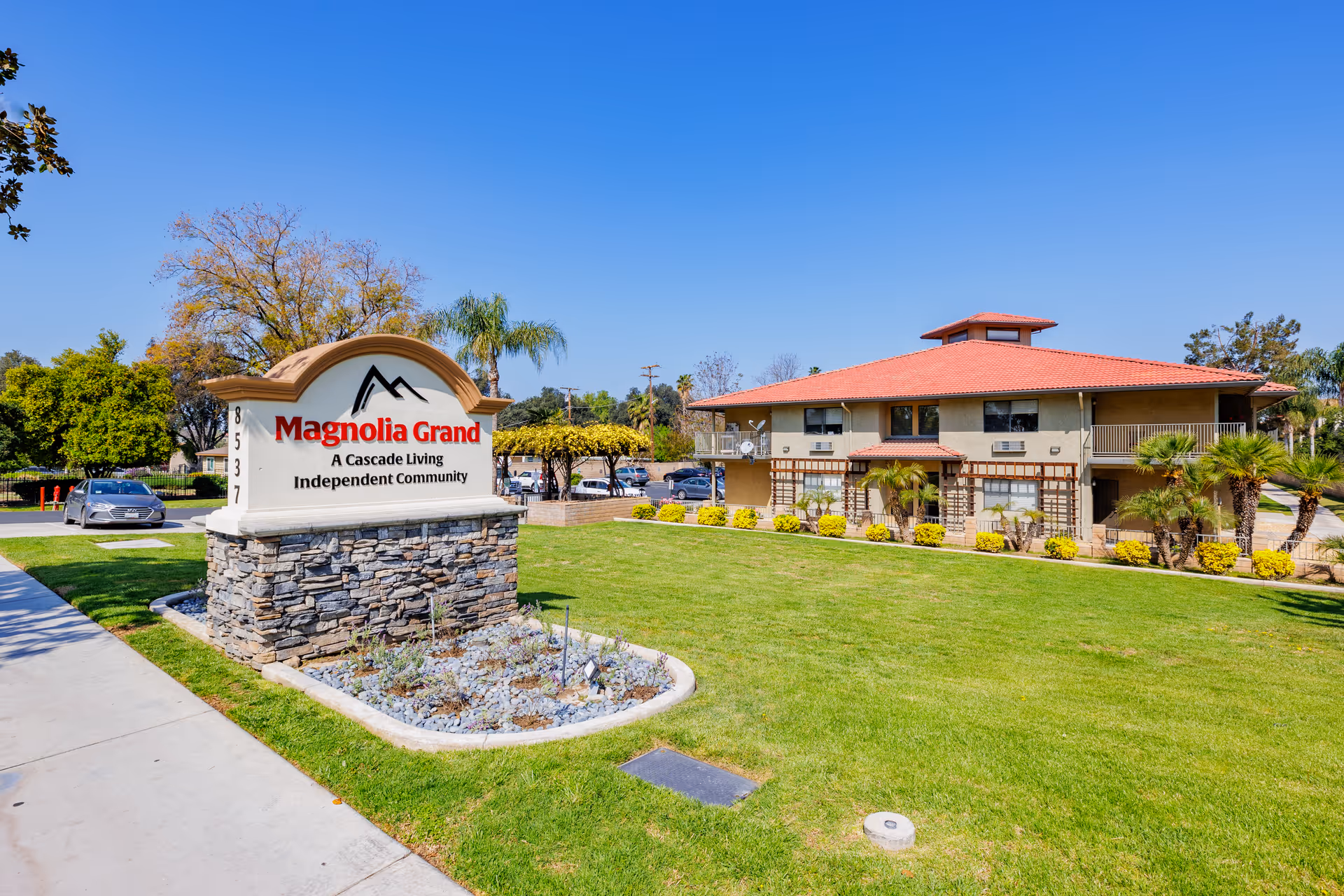 Entrance sign reading 'Magnolia Grand' in front of a two-story building with a manicured lawn under a clear blue sky.