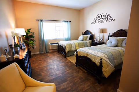 A well-lit bedroom with two twin beds featuring ornate dark wood headboards and matching bedding in soft green and beige tones. Between the beds is a nightstand with a lamp and decorative items. A window with light blue curtains allows natural light into the room. There is a wooden dresser with framed photos and a lamp on the left side, and a comfortable beige armchair in the foreground. The floor is covered with wood-like tiles.