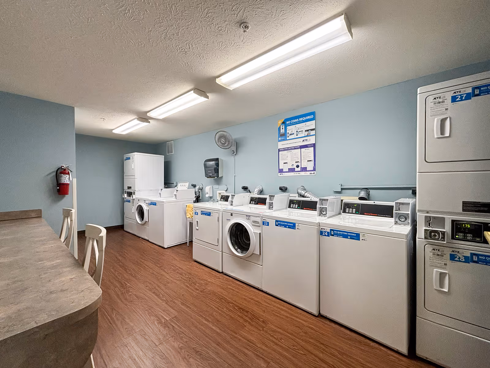 Laundry room with multiple washing machines and dryers lined up against a light blue wall. There is a countertop with chairs on the left side, wood flooring, and fluorescent ceiling lights. A fire extinguisher is mounted on the wall near the entrance.