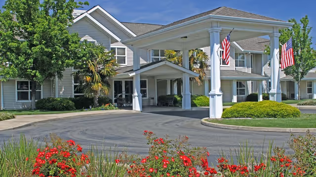 Front entrance of a senior living building with a covered porte-cochère, American flags, palm trees and red flowers in the foreground.