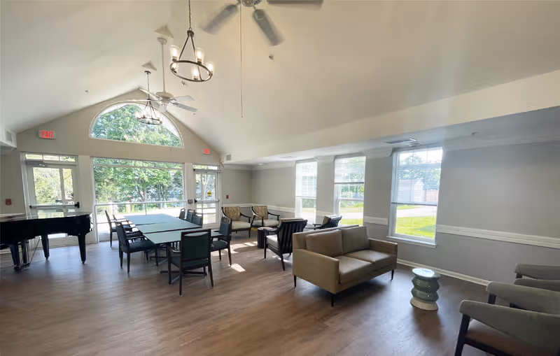 A bright and spacious common area in Quarry Station Seniors Apartments featuring large windows letting in natural light, multiple seating arrangements including chairs and sofas, a grand piano, and two ceiling fans with chandeliers hanging from a vaulted ceiling.