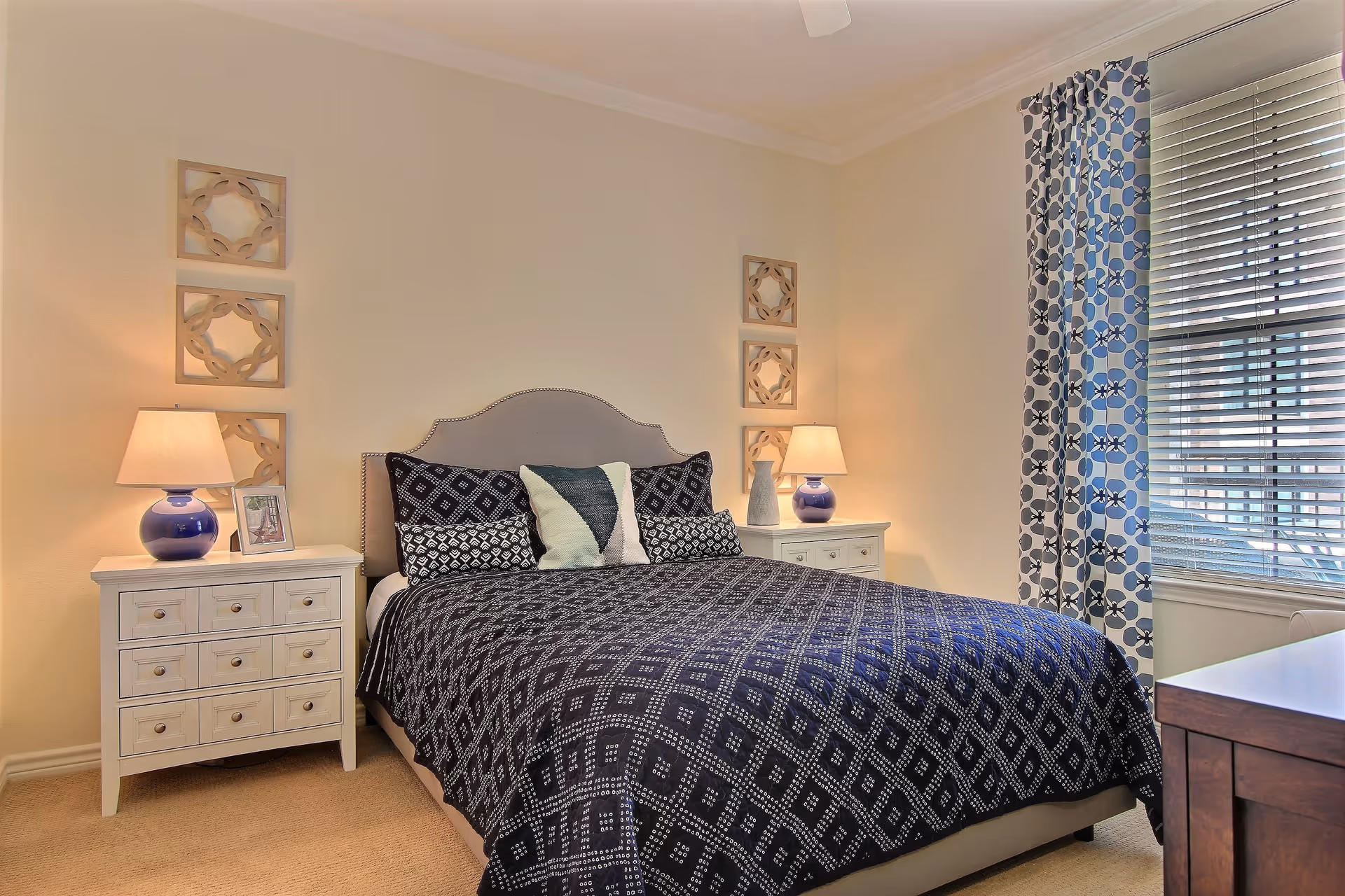 A bedroom with a bed covered in a dark blue patterned quilt and several pillows. There are two white nightstands on either side of the bed, each with a blue lamp and decorative items. The walls are light-colored with three decorative wall hangings above each nightstand. A window with blue and white patterned curtains and blinds is on the right side of the room.