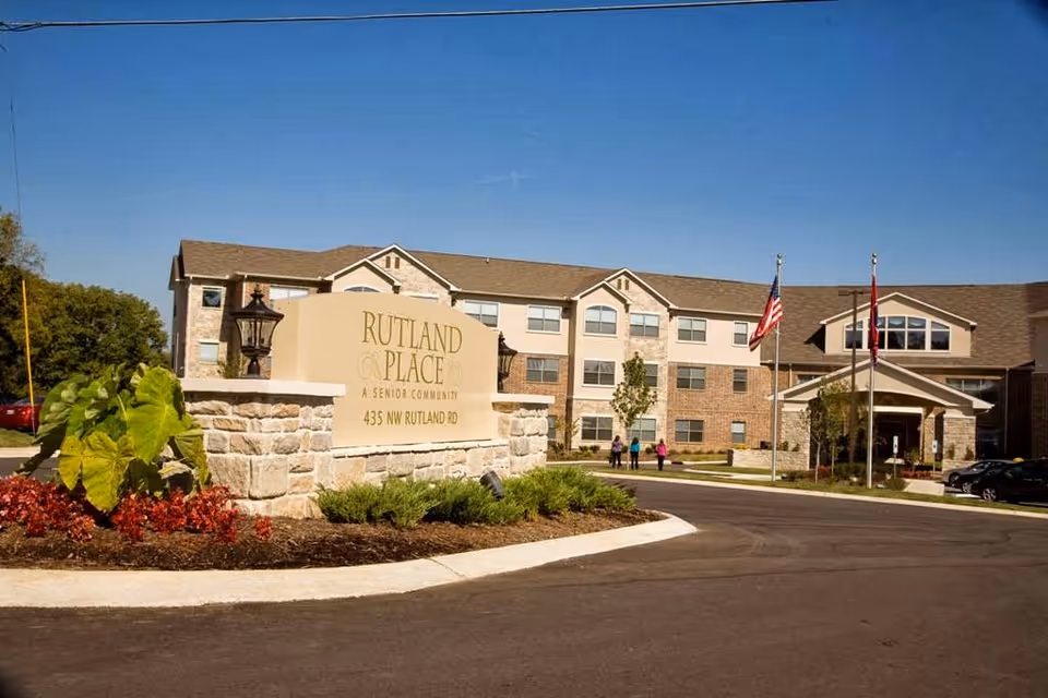 Front exterior of Rutland Place senior living building showing the entrance, flagpoles, landscaped sign, and driveway.
