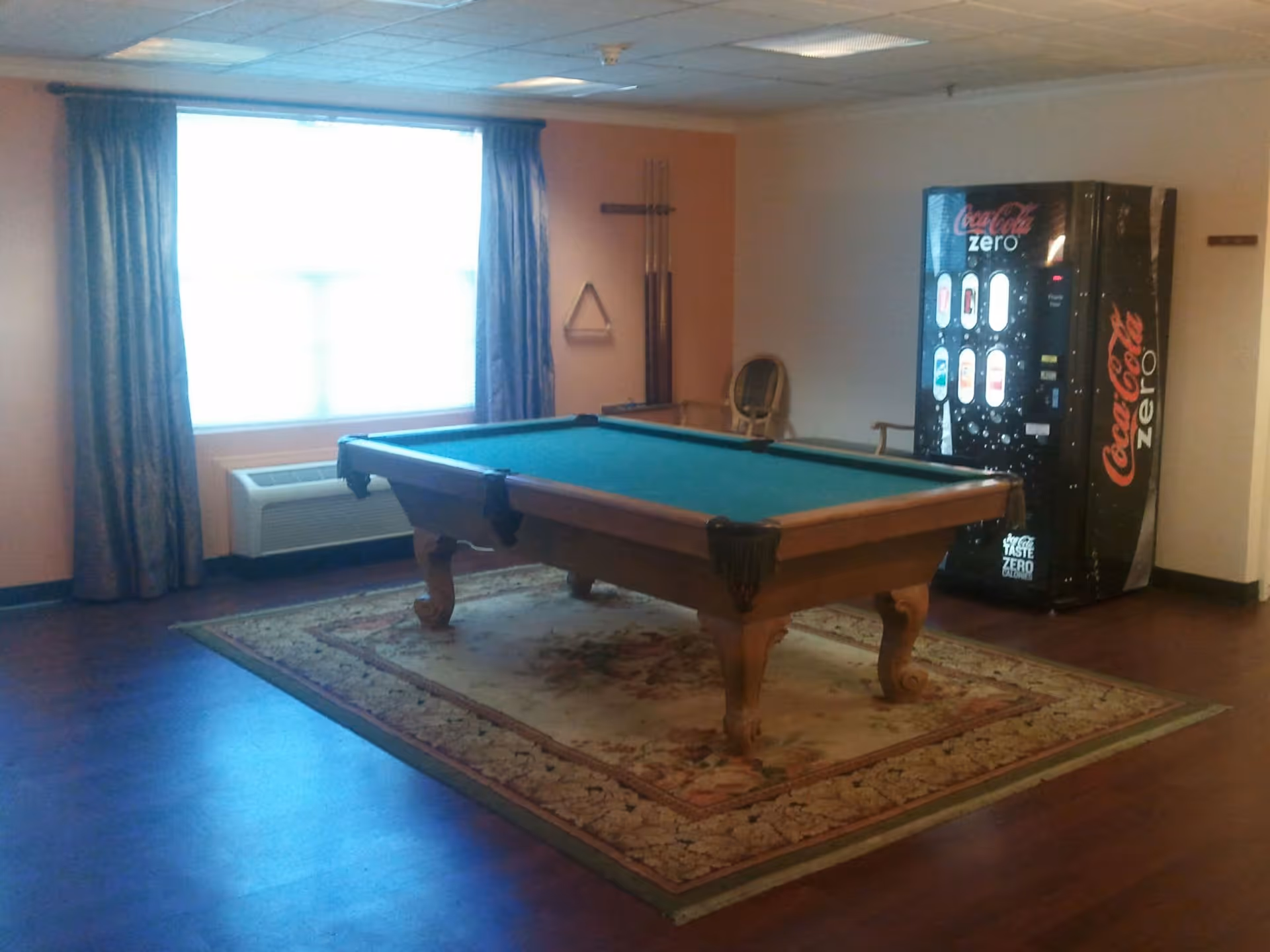 A room with a pool table placed on a decorative rug, a Coca-Cola Zero vending machine in the corner, a window with curtains, and a wall-mounted rack holding pool cues and a triangle rack.