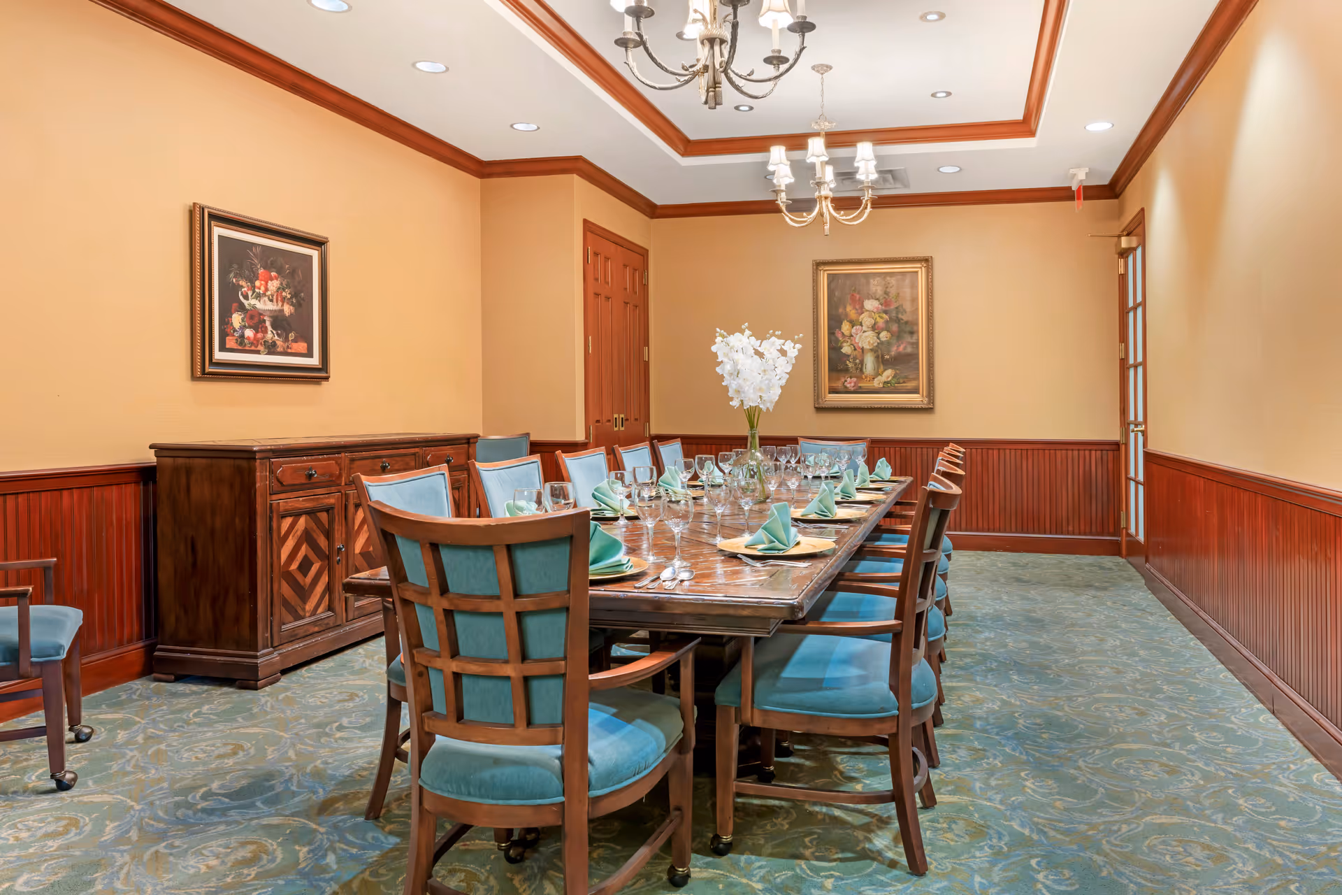A formal dining room with a long wooden table set for a meal, surrounded by wooden chairs with teal cushions. The table is decorated with folded green napkins, glassware, and a vase of white flowers in the center. The room has beige walls with wooden wainscoting, two framed floral paintings, a wooden sideboard, and two chandeliers hanging from a recessed ceiling.