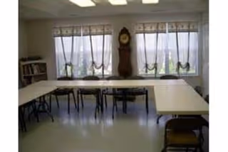 A room with several tables arranged in a U-shape and multiple chairs around them. There are three large windows with sheer curtains and valances, a grandfather clock between the windows, and a bookshelf in the corner. The floor is light-colored and the ceiling has fluorescent lights.