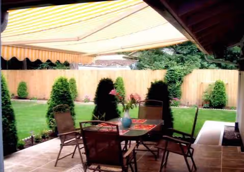 Covered outdoor patio with table and chairs, greenery, and a wooden privacy fence.