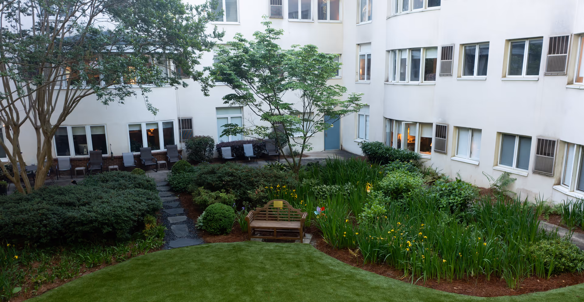 A landscaped courtyard garden area surrounded by a multi-story building with many windows. The garden features green shrubs, flowering plants, a small tree, a wooden bench, and a stone pathway. Several chairs are lined up against the building wall.