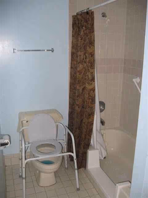 A bathroom with a shower-tub, curtain, and a toilet with a safety frame.