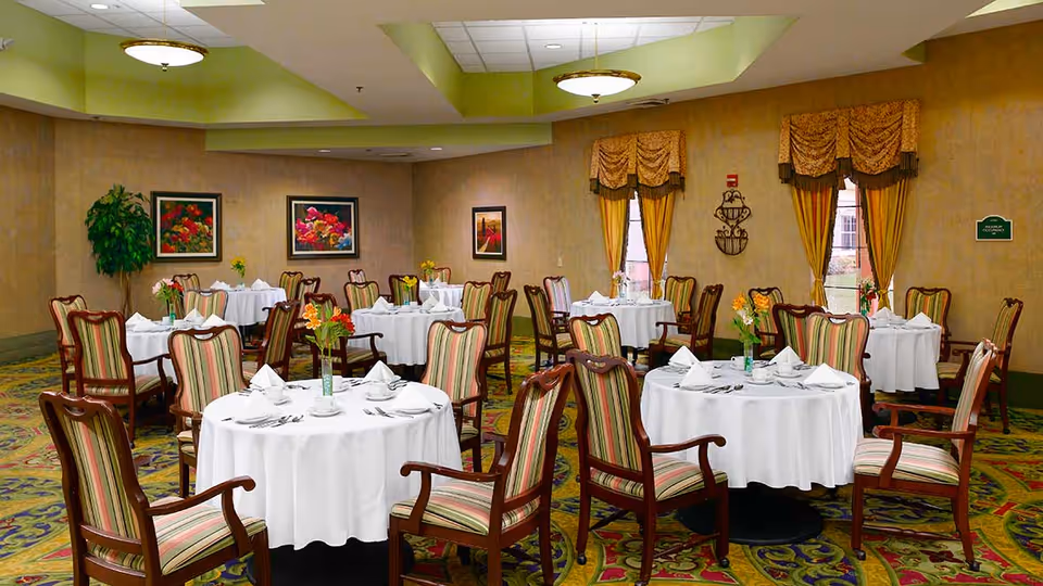 A dining room with multiple round tables covered with white tablecloths, each set with napkins, plates, and silverware. The chairs have wooden frames with striped upholstery. The room features patterned carpet, framed floral artwork on the walls, and windows with gold curtains.