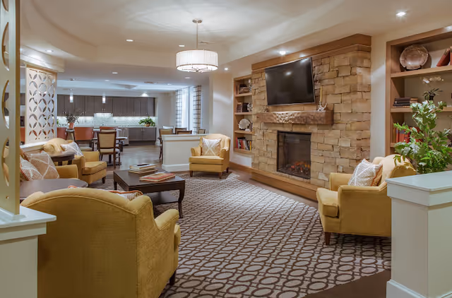 Comfortable communal lounge with yellow armchairs, a stone fireplace topped by a TV, and a dining area and kitchen in the background.