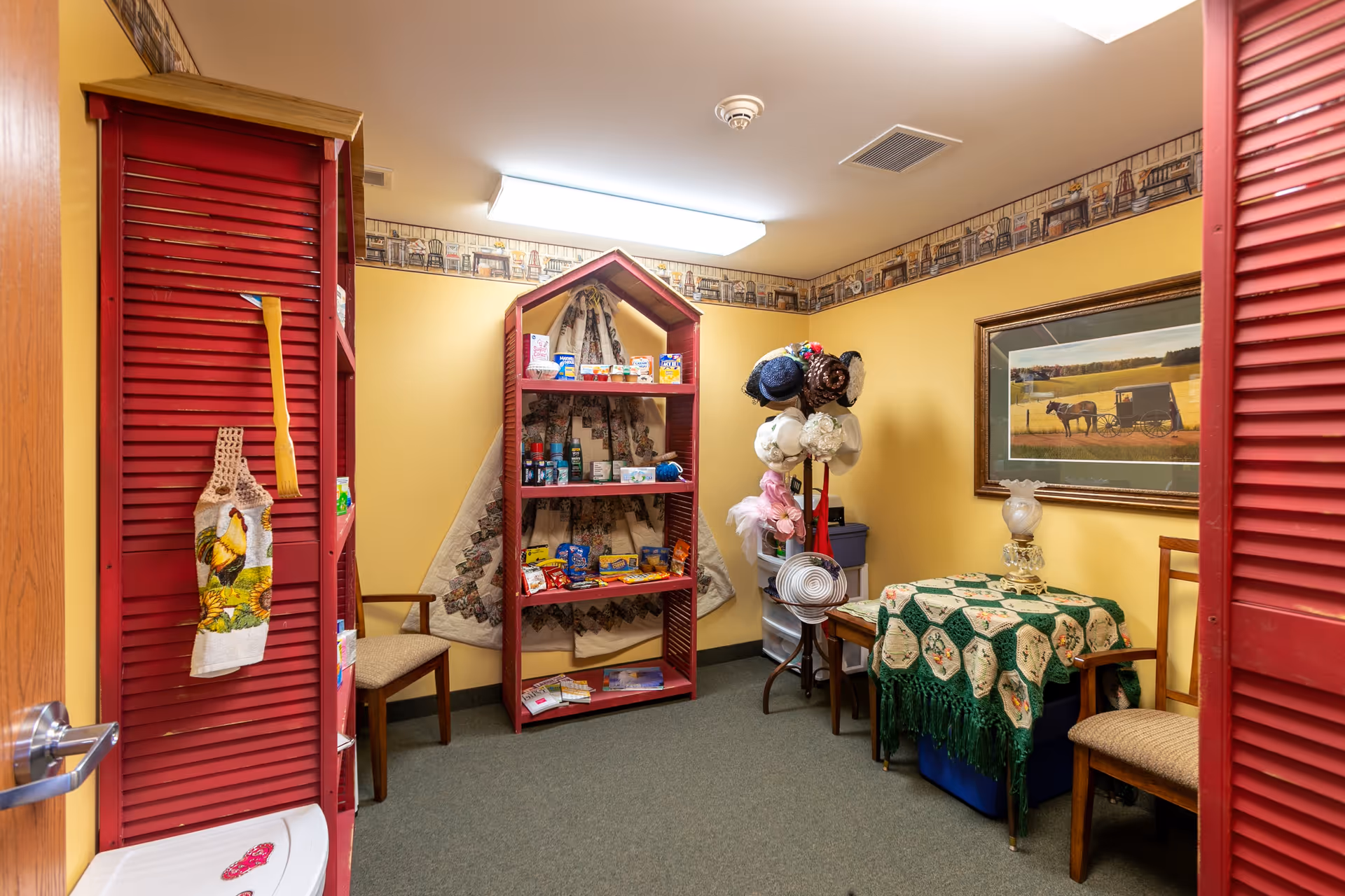 A small room with yellow walls and green carpet featuring red wooden shelves stocked with various items, a rack holding multiple hats, two wooden chairs, a small table covered with a green and beige crocheted tablecloth, and a framed picture of a horse and buggy on the wall.