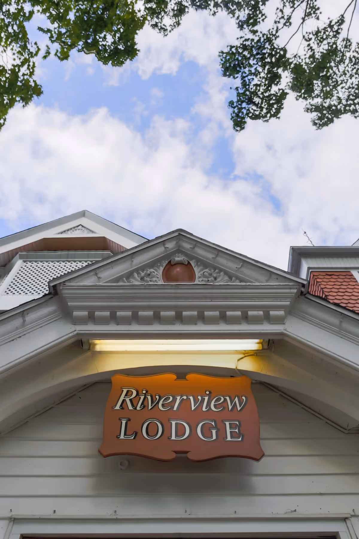 Upward view of the entrance of a building with a decorative triangular pediment and a sign that reads 'Riverview Lodge' hanging below it. The sky is partly cloudy with tree branches framing the top of the image.