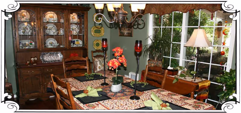 A dining room with a wooden dining table covered with a patterned tablecloth and set with black placemats and green folded napkins. The table has a centerpiece with red flowers and two tall red candle holders. Behind the table is a wooden china cabinet displaying plates and glassware. To the right, there is a large window with a view of greenery outside and a table lamp on a side table with plants around it.