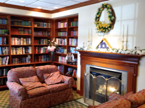Cozy living room area with floral patterned armchairs and a loveseat in front of a wooden fireplace with a lit fire. Above the fireplace is a decorative wreath and a folded American flag displayed in a triangular case. To the left, there is a large wooden bookshelf filled with books and decorative items.