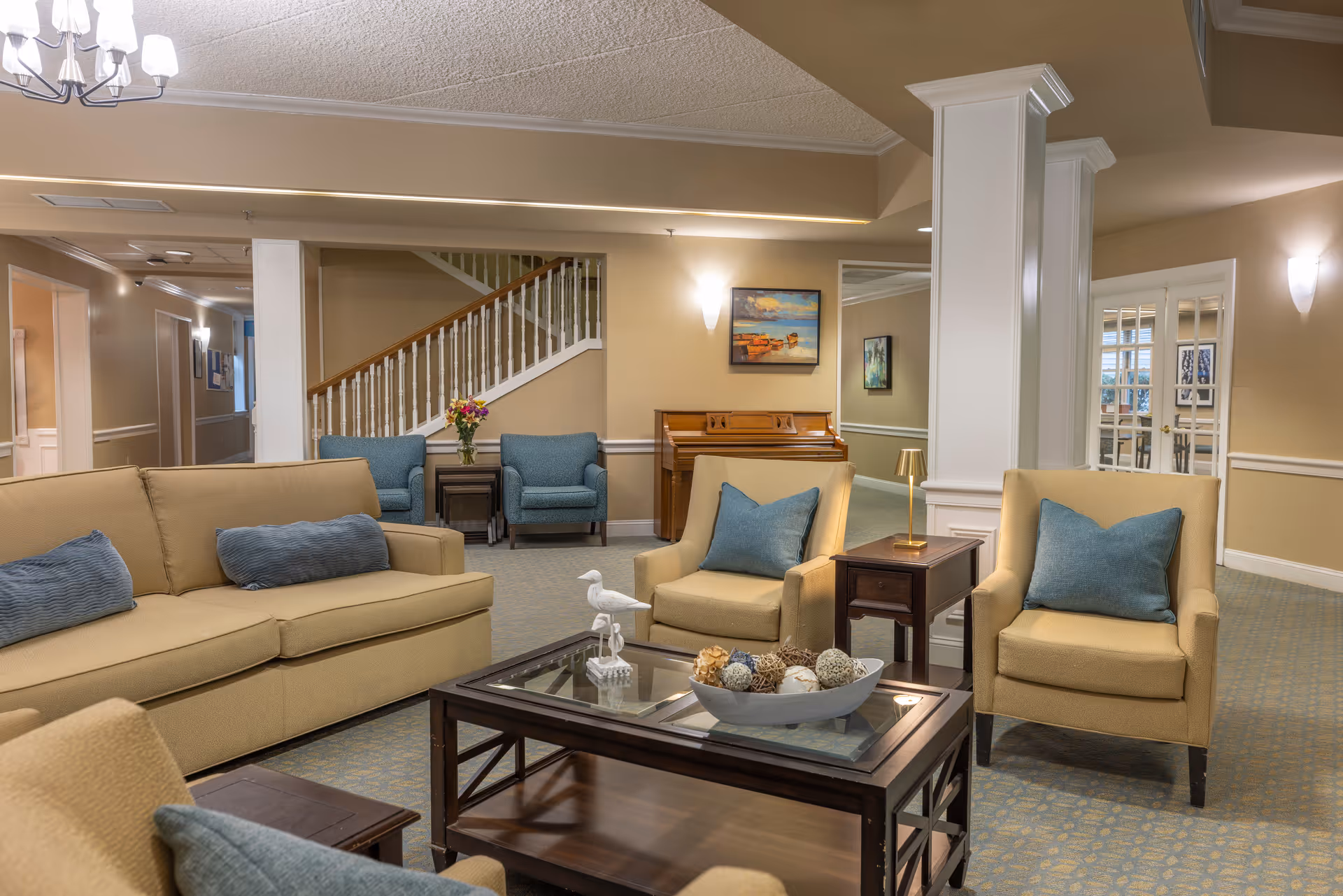 A cozy living room area in an assisted living facility featuring beige sofas and armchairs with blue cushions arranged around a glass-top coffee table decorated with a bowl of decorative spheres and a small bird figurine. In the background, there is a wooden piano, a staircase with white railings, and a hallway. The walls are painted beige with white trim, and the room is softly lit with wall sconces and a chandelier.