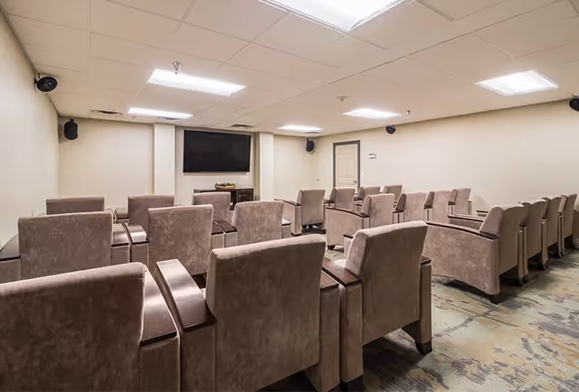 A small theater-style room with multiple rows of comfortable beige armchairs facing a large flat-screen TV mounted on the wall. The room has a beige carpet with a subtle pattern, beige walls, and a white drop ceiling with fluorescent lighting. There are speakers mounted in the corners of the room.