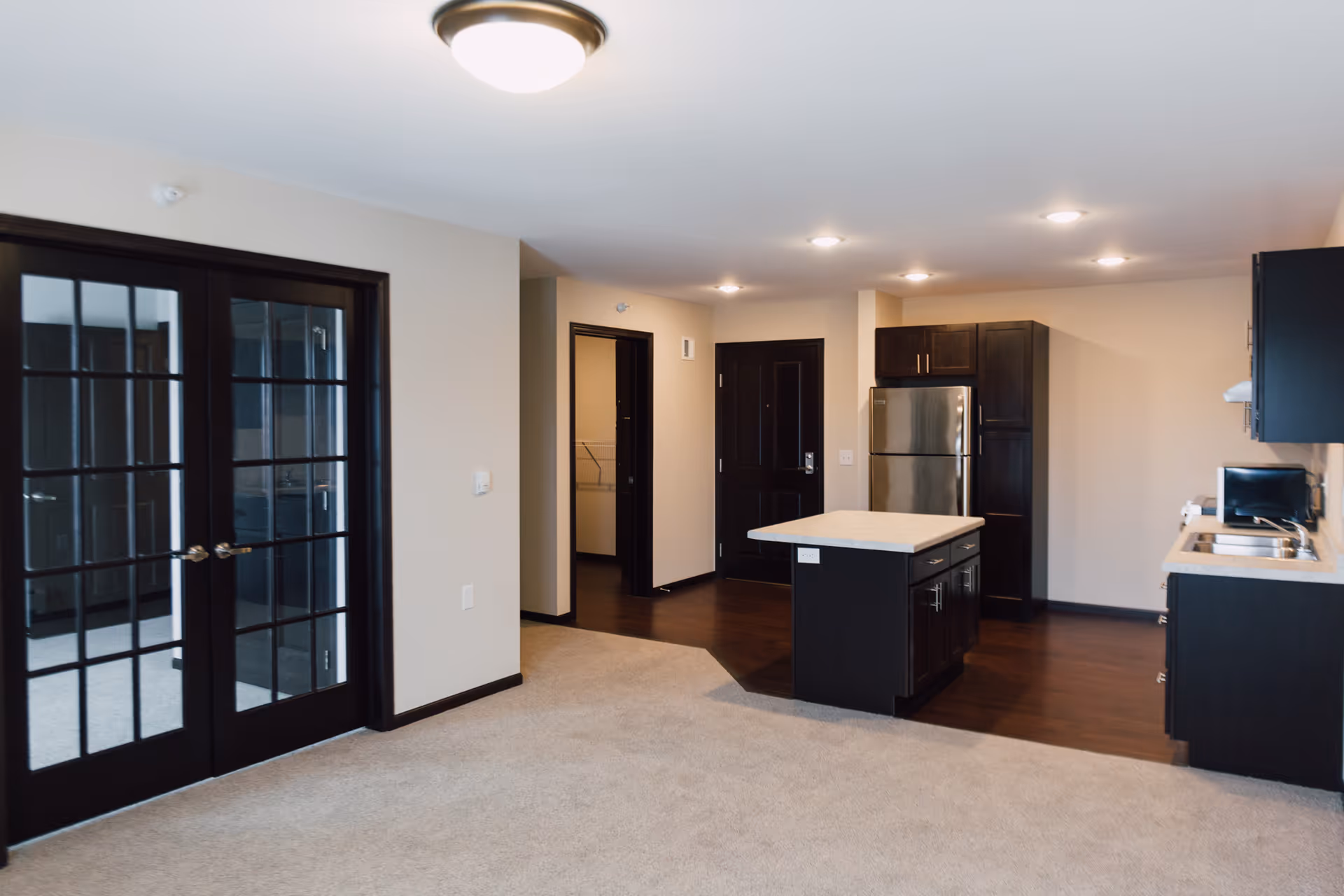 Interior view of a modern apartment unit at Pelican Landing featuring a kitchen with dark wood cabinets, a stainless steel refrigerator, a kitchen island with a light countertop, and a living area with beige carpet. There are double glass doors on the left side and a dark wood entrance door in the background.