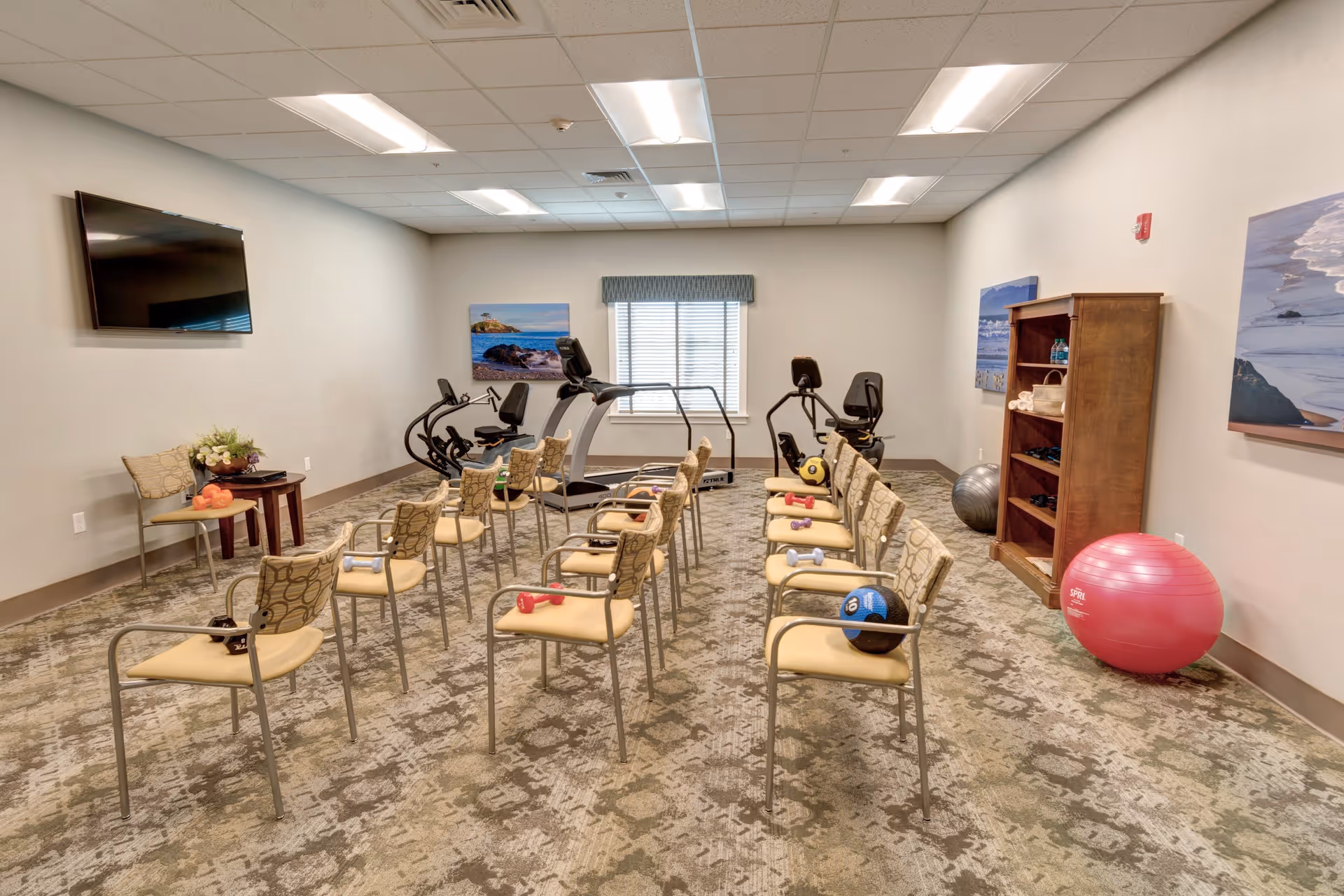 A fitness room with two rows of chairs, each holding small exercise weights and balls. The room has exercise equipment including a treadmill and stationary bikes near a window. There is a wooden shelf with towels and water bottles, a large pink exercise ball, and framed pictures of coastal scenes on the walls. A flat-screen TV is mounted on one wall.