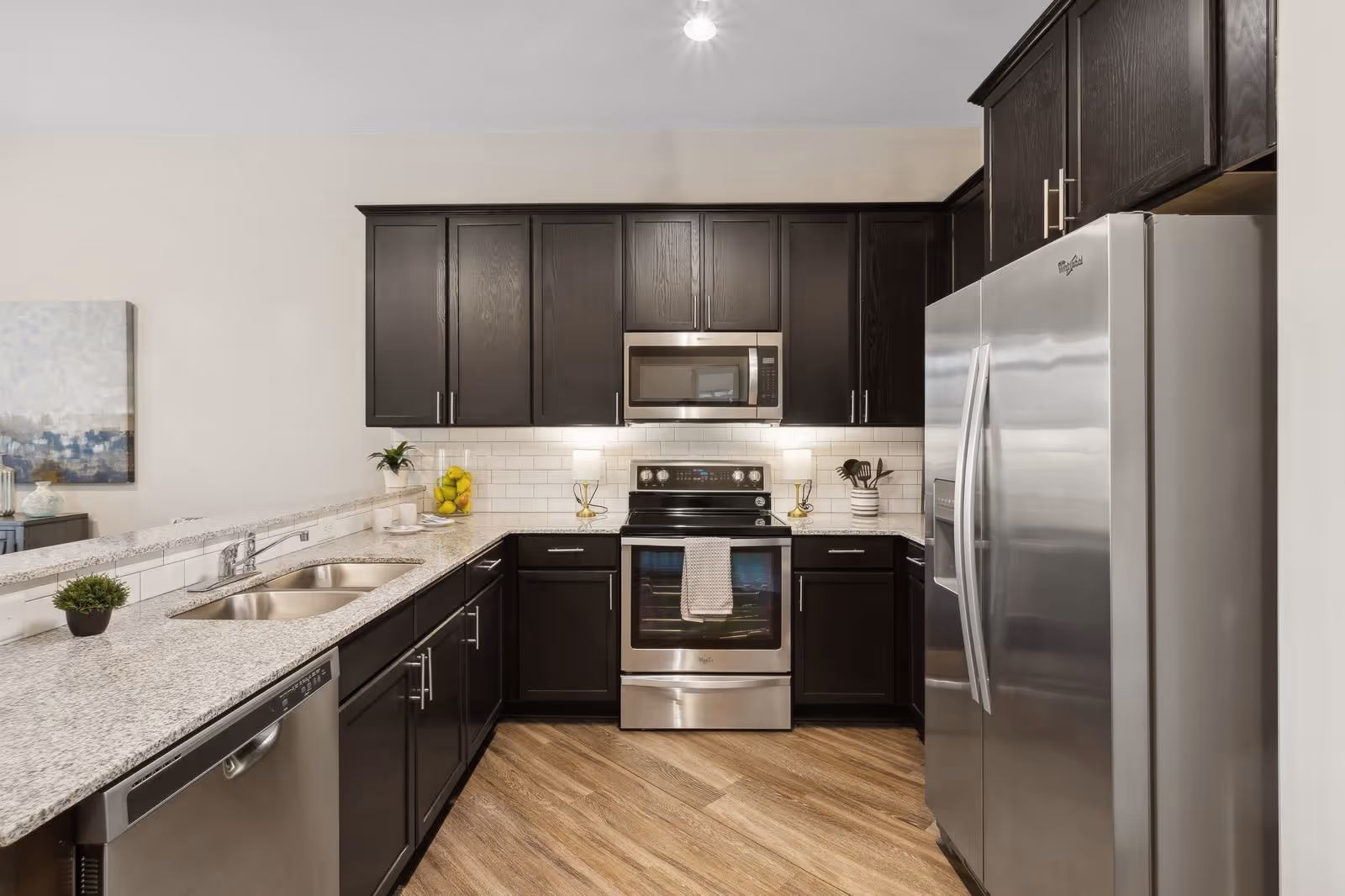 Modern kitchen with dark wood cabinets, stainless steel appliances including a refrigerator, oven, microwave, and dishwasher. The kitchen has a granite countertop with a double sink, white subway tile backsplash, and wood-patterned flooring. There are decorative items such as a small plant, a bowl of lemons, and kitchen utensils on the counter.