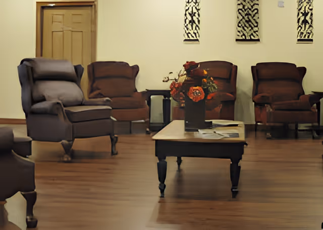 A communal seating area with upholstered armchairs, a central coffee table topped with a floral arrangement, and wood flooring.