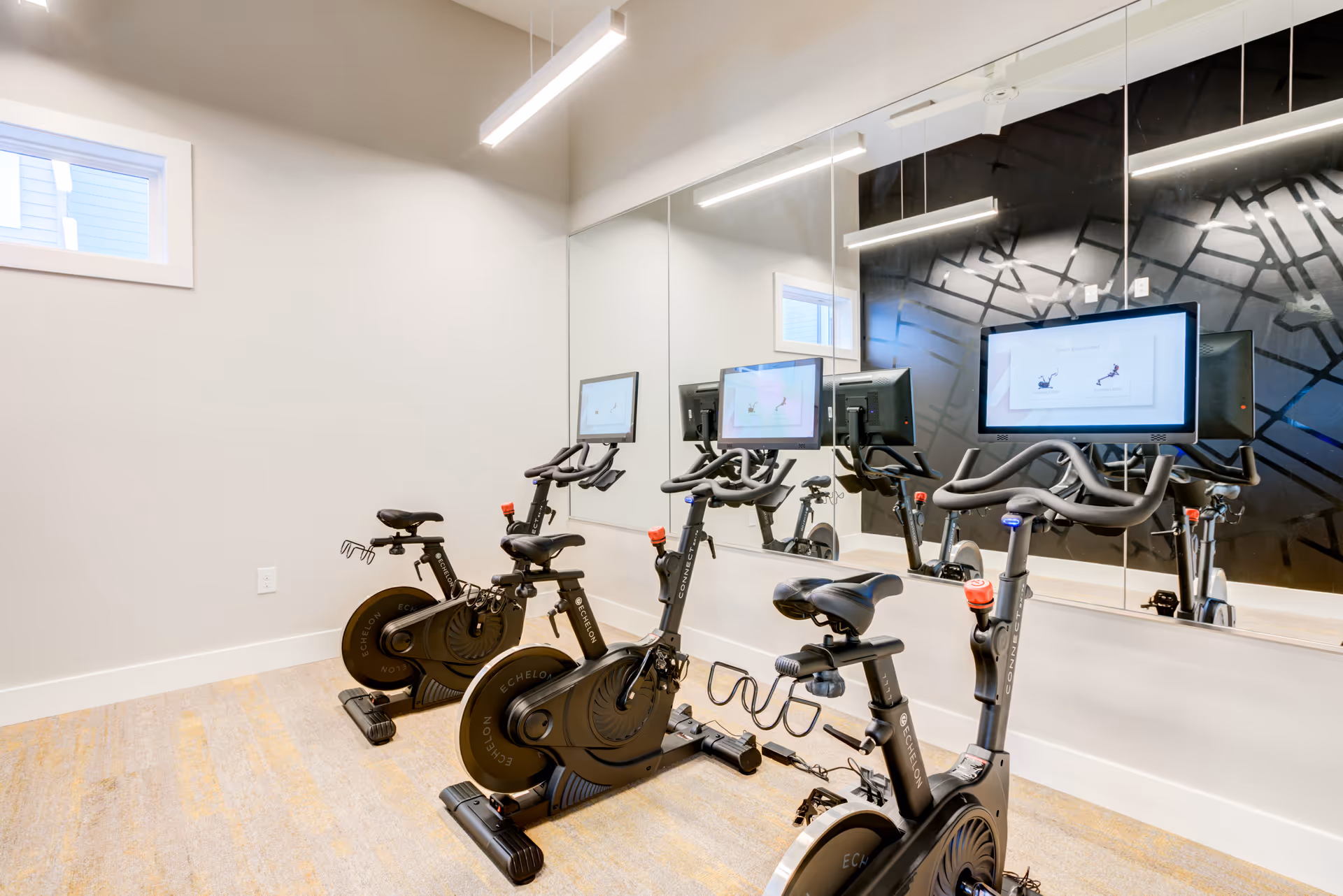 Three stationary exercise bikes with individual screens arranged in front of a mirrored wall in a bright fitness room.