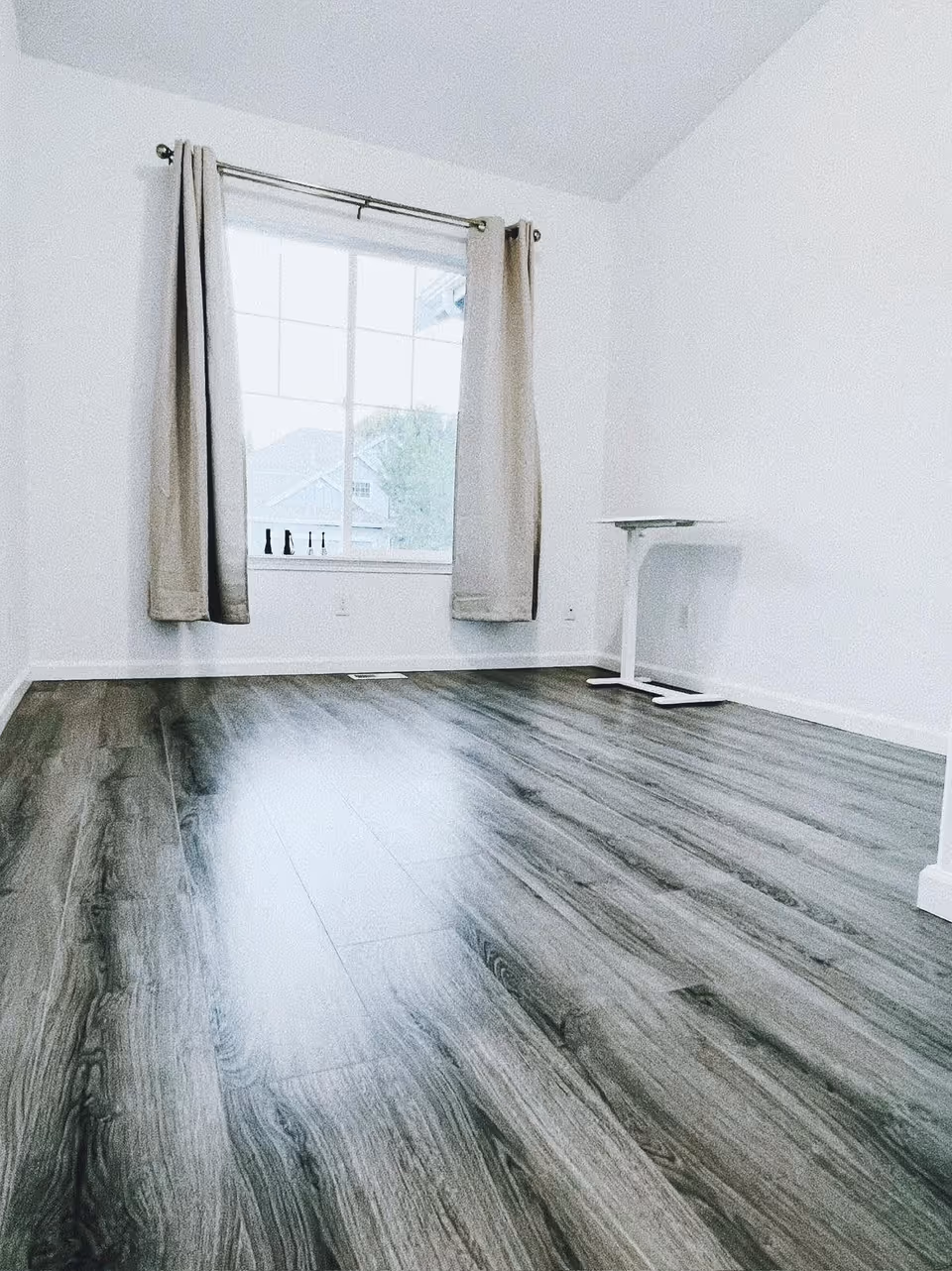Empty room with gray wood flooring, white walls, a large window with beige curtains, and a small white table in the corner.
