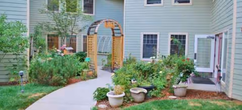 Outdoor courtyard with a curved path, potted plants, and a wooden arch beside a green building.