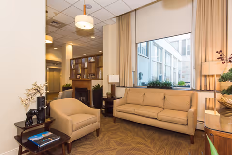 A cozy interior common area with a beige sofa and matching armchair, a small side table with decorative items including a black elephant figurine, a fireplace with shelves above it, large windows with beige curtains, and warm lighting from ceiling fixtures and lamps.
