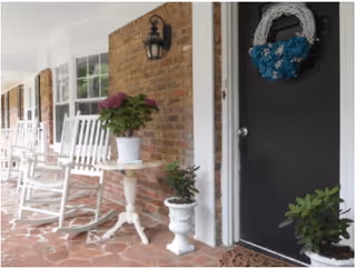 A covered porch area with white rocking chairs and small tables holding potted plants. The porch has a brick wall with a black door decorated with a blue and white wreath. There are additional potted plants on the floor near the door.