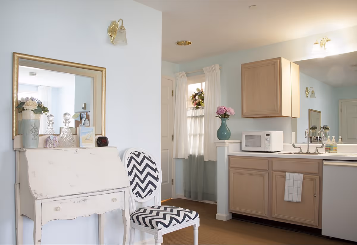 A cozy interior space featuring a small kitchen area with light wood cabinets, a white microwave, a sink with a faucet, and a countertop with soap dispensers. Adjacent to the kitchen is a white distressed writing desk with decorative items including a vase with flowers, crystal decanters, and a small clock. A white chair with a black and white chevron patterned cushion sits next to the desk. The room has light blue walls, a large mirror above the desk, a window with white curtains, and soft lighting fixtures on the walls.
