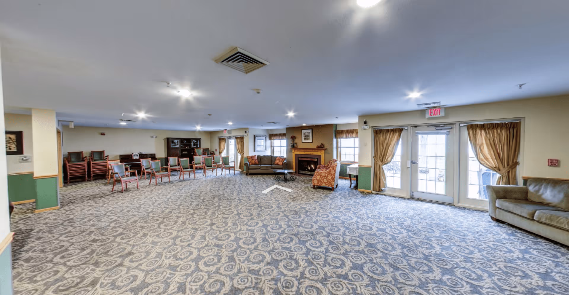 Spacious senior living common room with patterned carpet, multiple chairs and sofas, a fireplace, and glass exit doors.