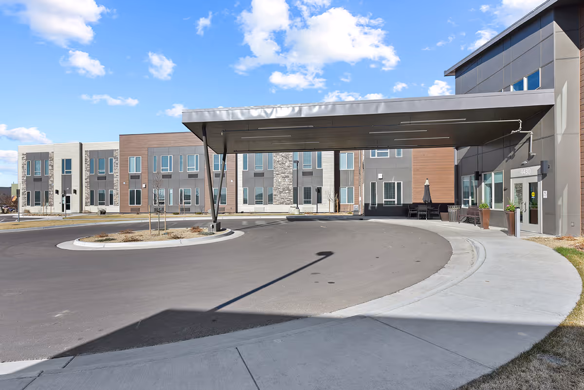 Exterior view of The Lodge at Greeley senior living facility showing a modern building with a covered driveway entrance, multiple windows, and a clear blue sky with some clouds.