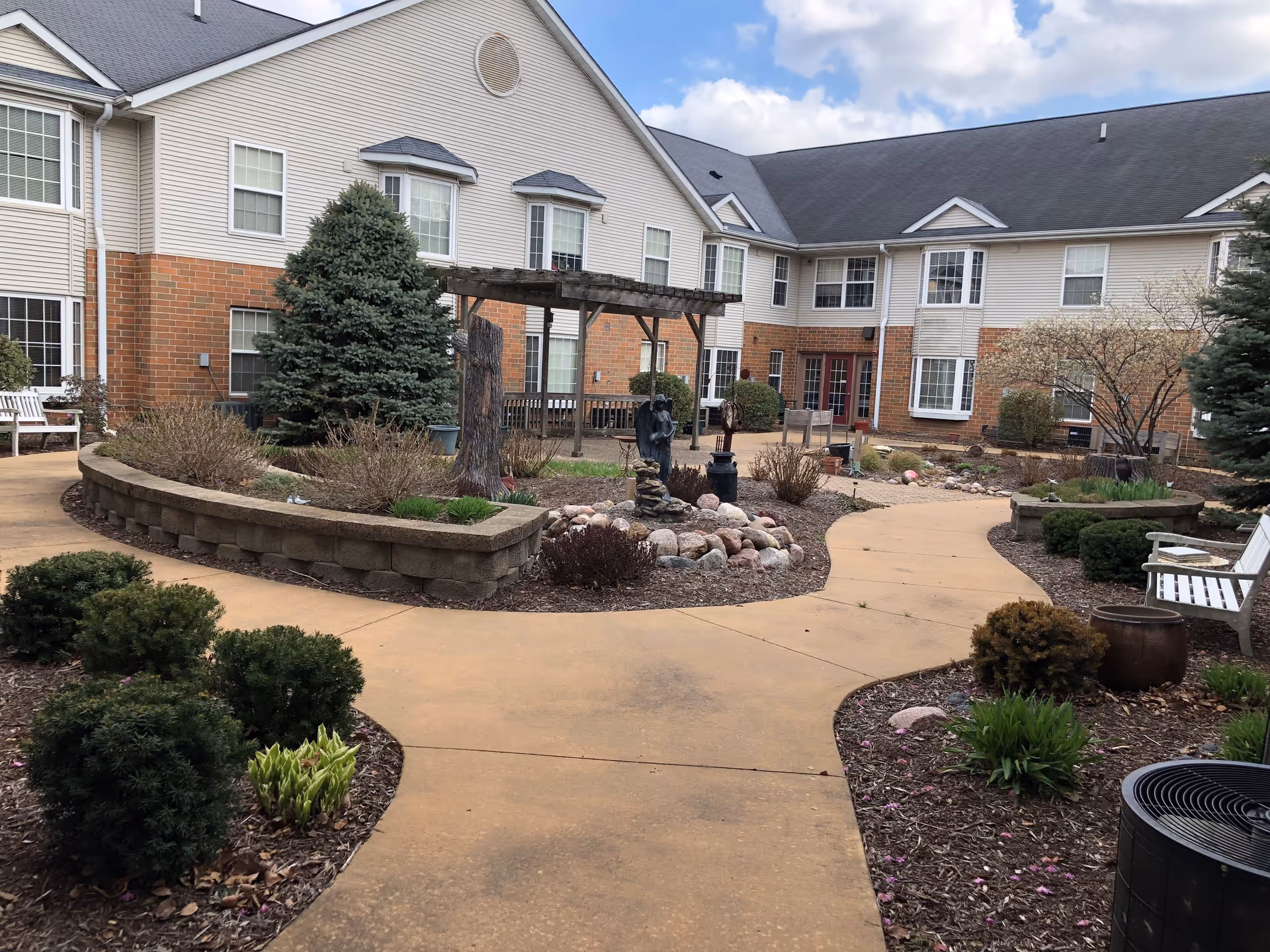 Outdoor courtyard area of a senior living facility with paved walkways, landscaped garden beds, bushes, small trees, benches, and a wooden pergola structure. The building surrounding the courtyard is two stories with beige siding and brick accents under a cloudy sky.