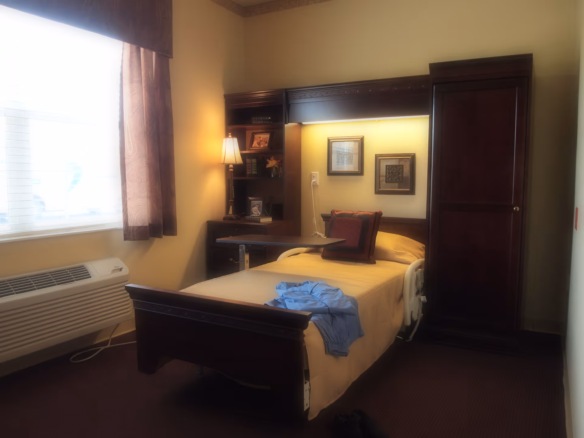 A warm, softly lit senior bedroom with a single hospital-style bed, wooden headboard and shelving, lamp, window with curtains, and a wardrobe.