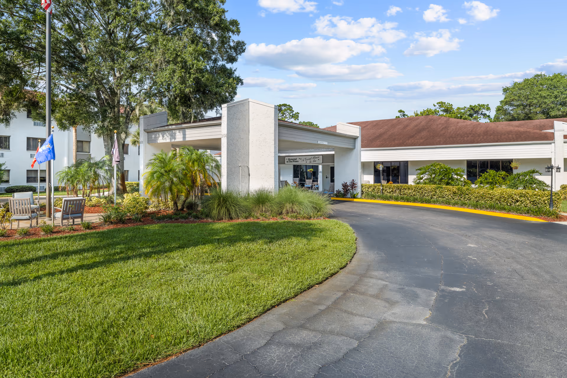 Front entrance of a senior living facility with a covered drop-off, curved driveway, flags, and landscaped grounds.