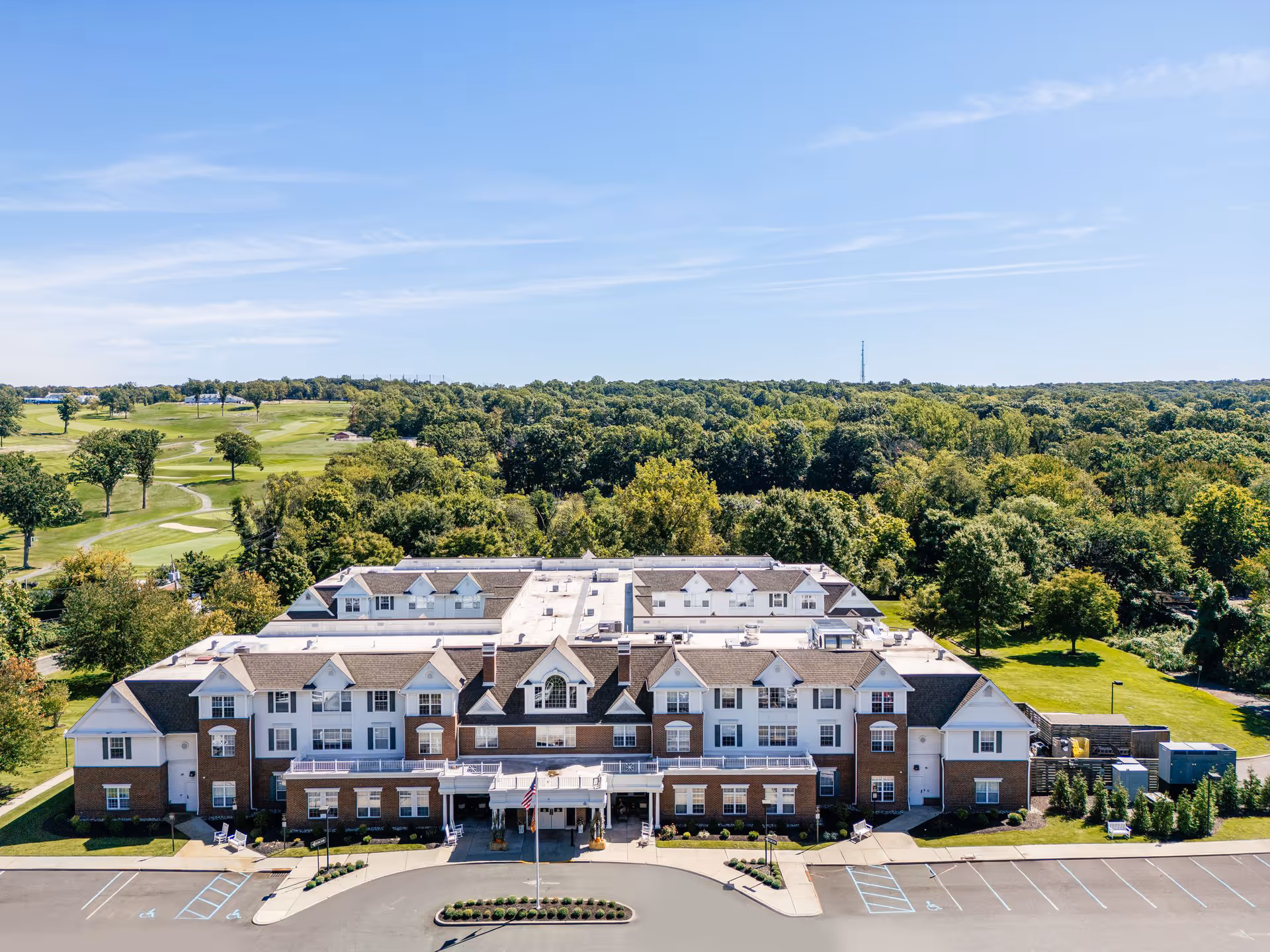 Aerial view of the front of a large three-story senior living building with a parking lot, surrounding trees, and a golf course in the background.