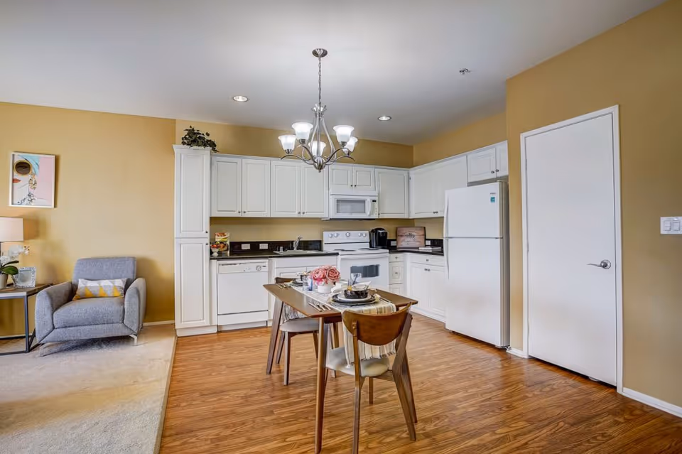 A bright kitchen and dining area with white cabinets, a white refrigerator, stove, microwave, and dishwasher. A wooden dining table with four chairs is set with plates, cups, and a floral centerpiece. To the left, there is a gray armchair with a yellow and white pillow next to a side table with a lamp and a small plant. The walls are painted a warm beige color and the floor is wood with a carpeted area near the armchair.