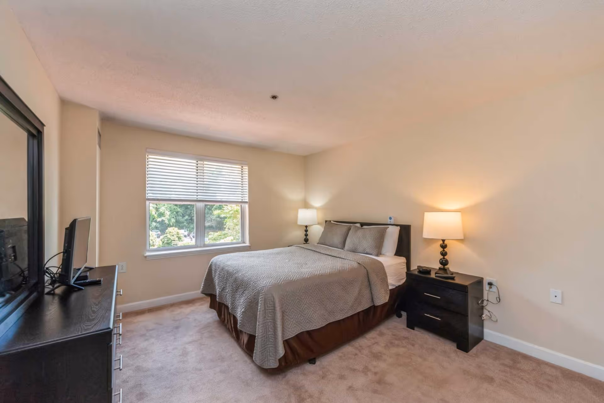 A bedroom with a neatly made bed covered with a gray quilt and matching pillows. There is a black nightstand with a lamp on the right side of the bed. A window with white blinds lets in natural light, and a black dresser with a TV on top is positioned on the left side of the room. The walls are painted beige and the floor is carpeted.