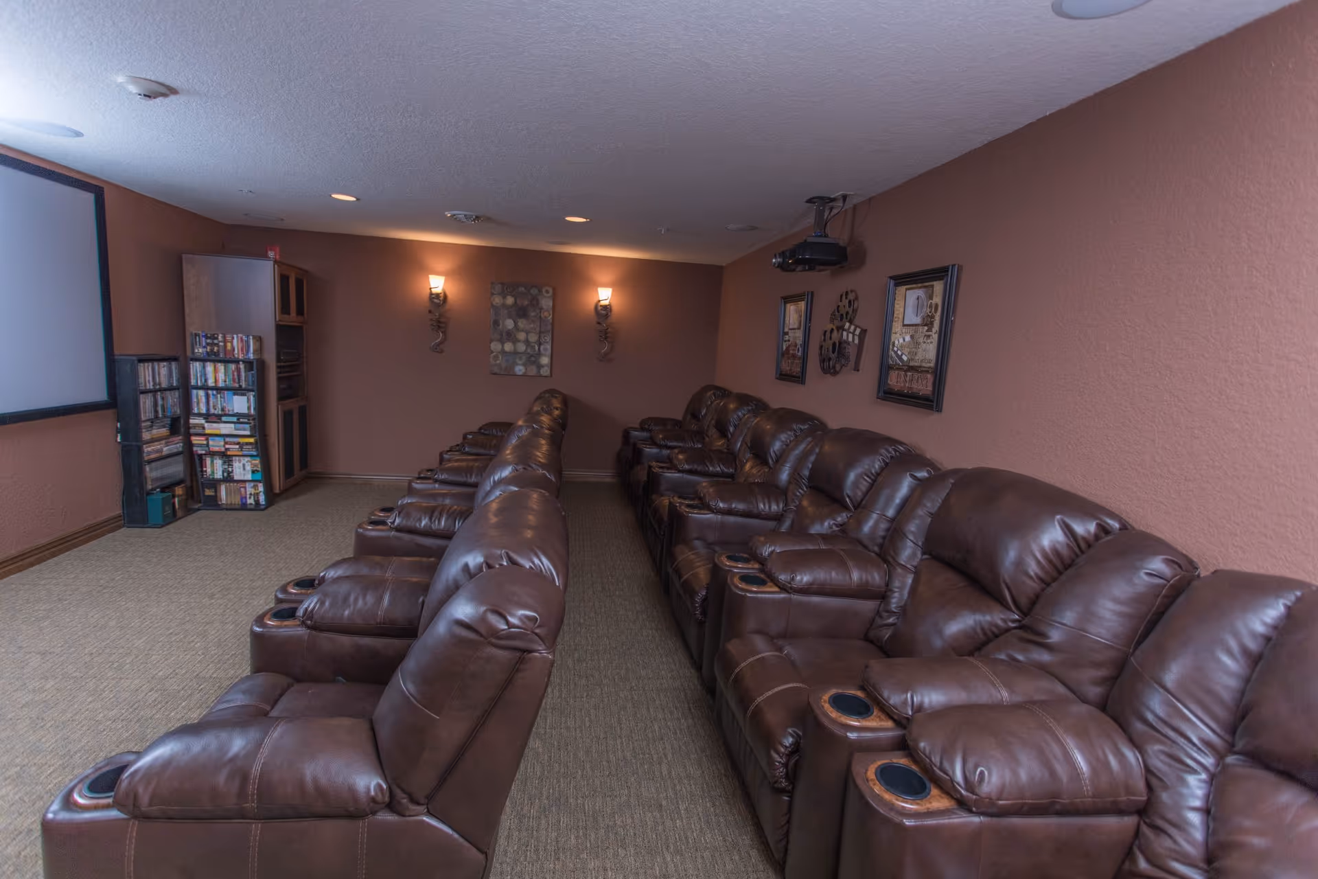 Rows of brown leather recliner chairs facing a projection screen in a cozy media/theater room with wall sconces and shelving of DVDs.