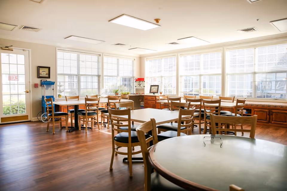 A bright dining room with multiple wooden tables and chairs arranged neatly. Large windows with white blinds allow natural light to fill the room. There is a popcorn machine and some plants on a counter near the windows. The floor is wooden, and the walls are painted light beige.