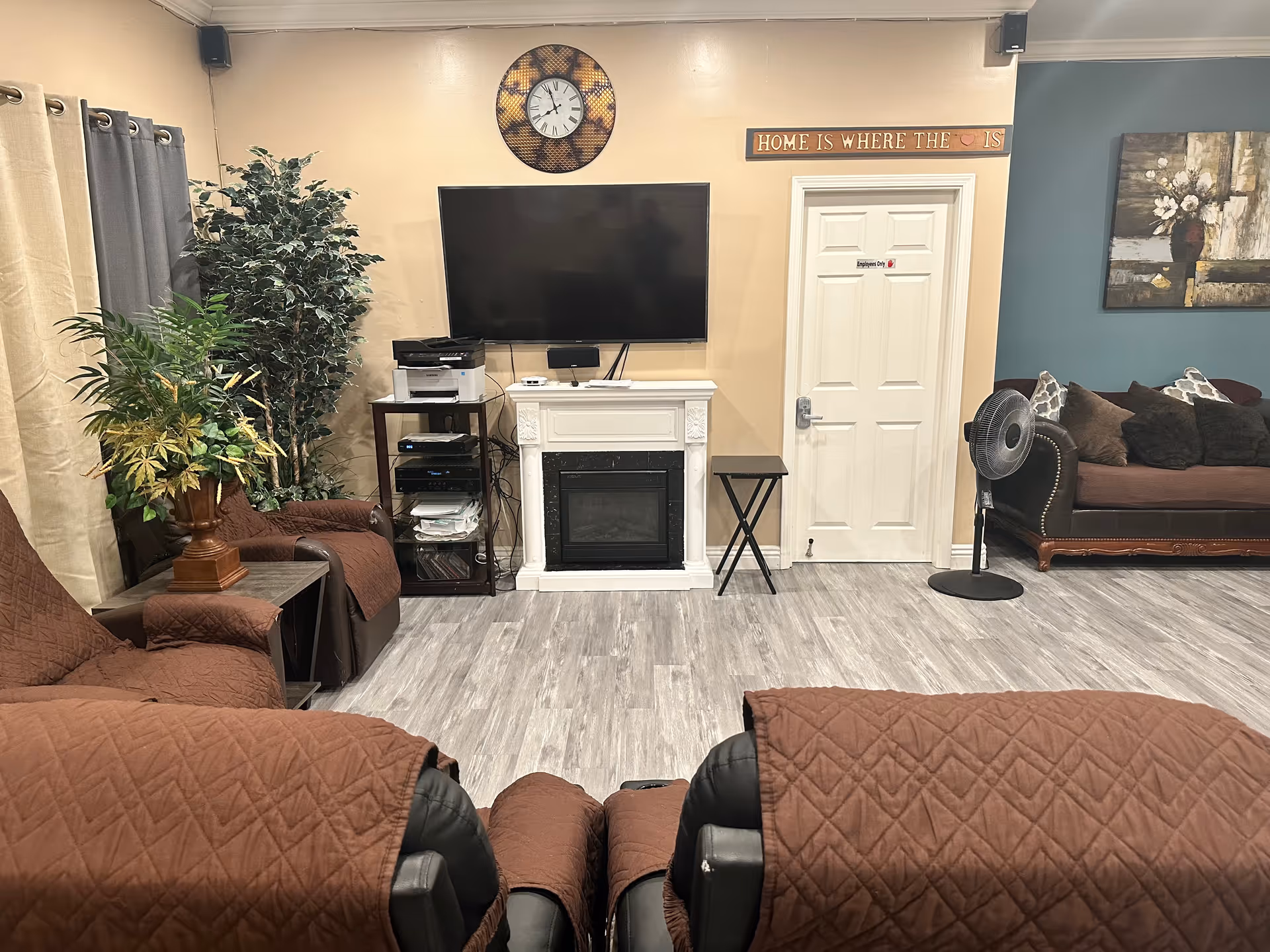 A cozy living room with brown recliner chairs covered with brown quilted covers, a small table with a potted plant, a large flat-screen TV mounted on a beige wall above a white electric fireplace, and a clock above the TV. To the right is a white door with a sign that reads 'Employees Only' and a wooden sign above it that says 'HOME IS WHERE THE ♥ IS'. There is a dark brown couch with multiple pillows against a blue accent wall, a standing fan, and a piece of floral artwork on the wall.